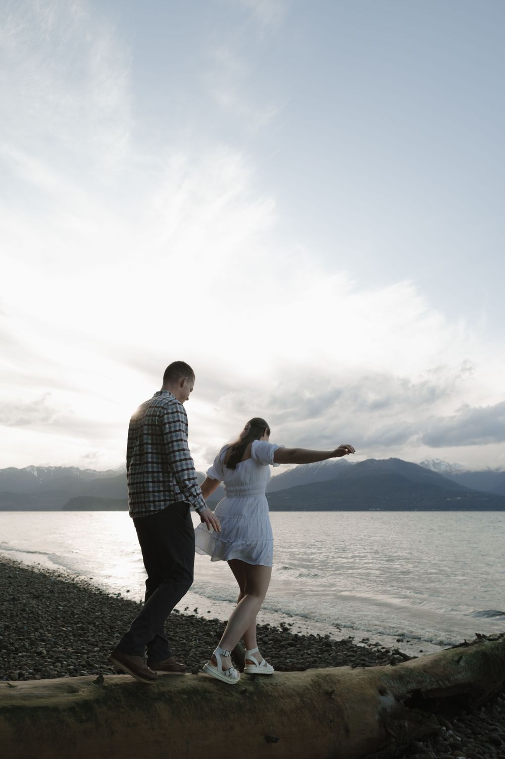 Two people balancing on driftwood at the edge of a scenic mountain lake.