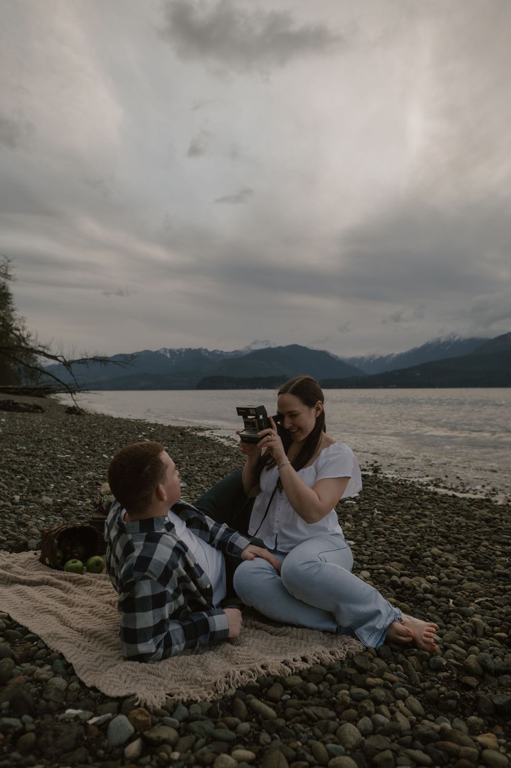 Relaxed picnic scene on a blanket by the lake as someone takes photos.