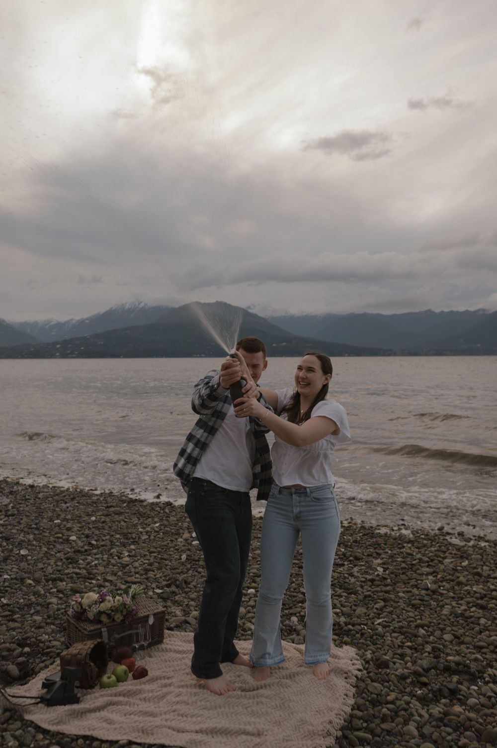 Happy couple celebrating with champagne on a rocky beach overlooking mountains.