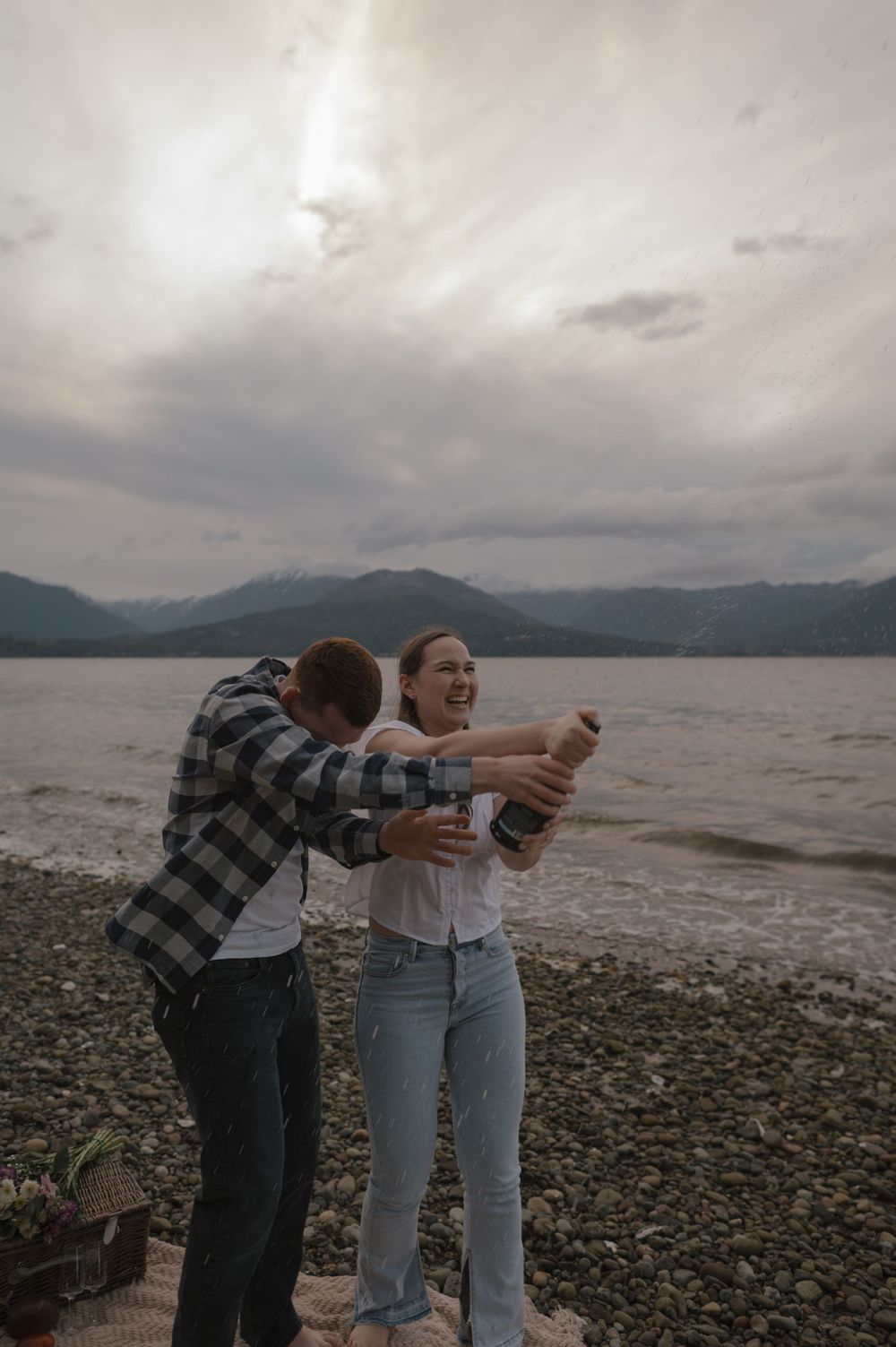 Joyful moment as couple pops champagne on a rocky beach with mountain views.