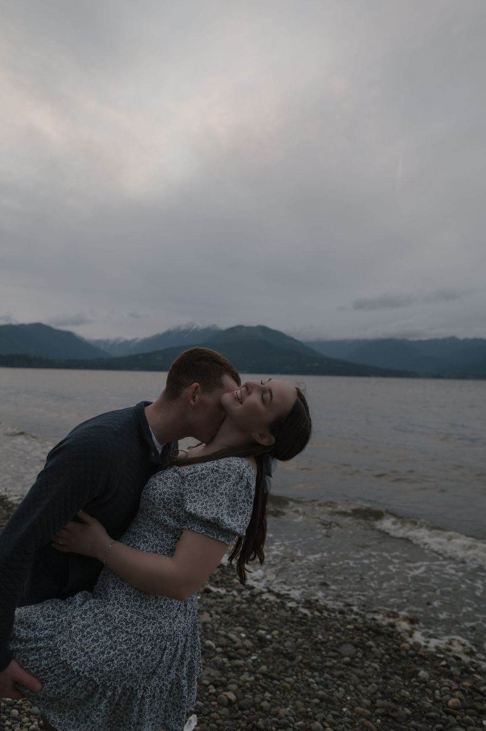 A romantic couple embraces on a rocky beach at dusk with mountains and cloudy skies in the background.