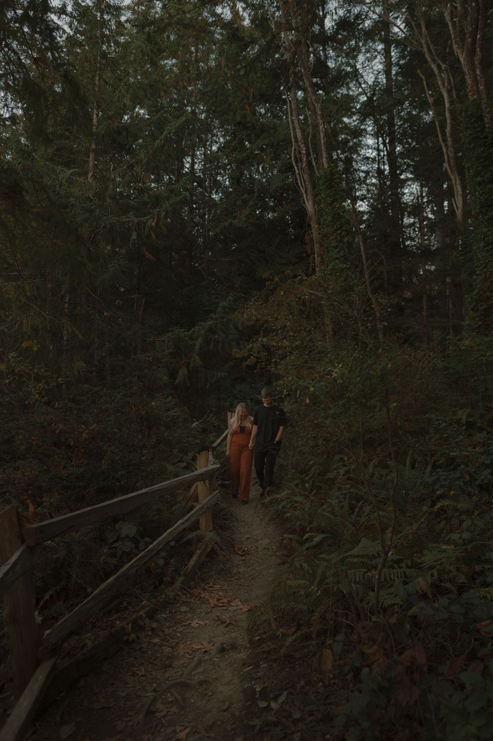 Couple walking along a forested trail with wooden railings at dusk.