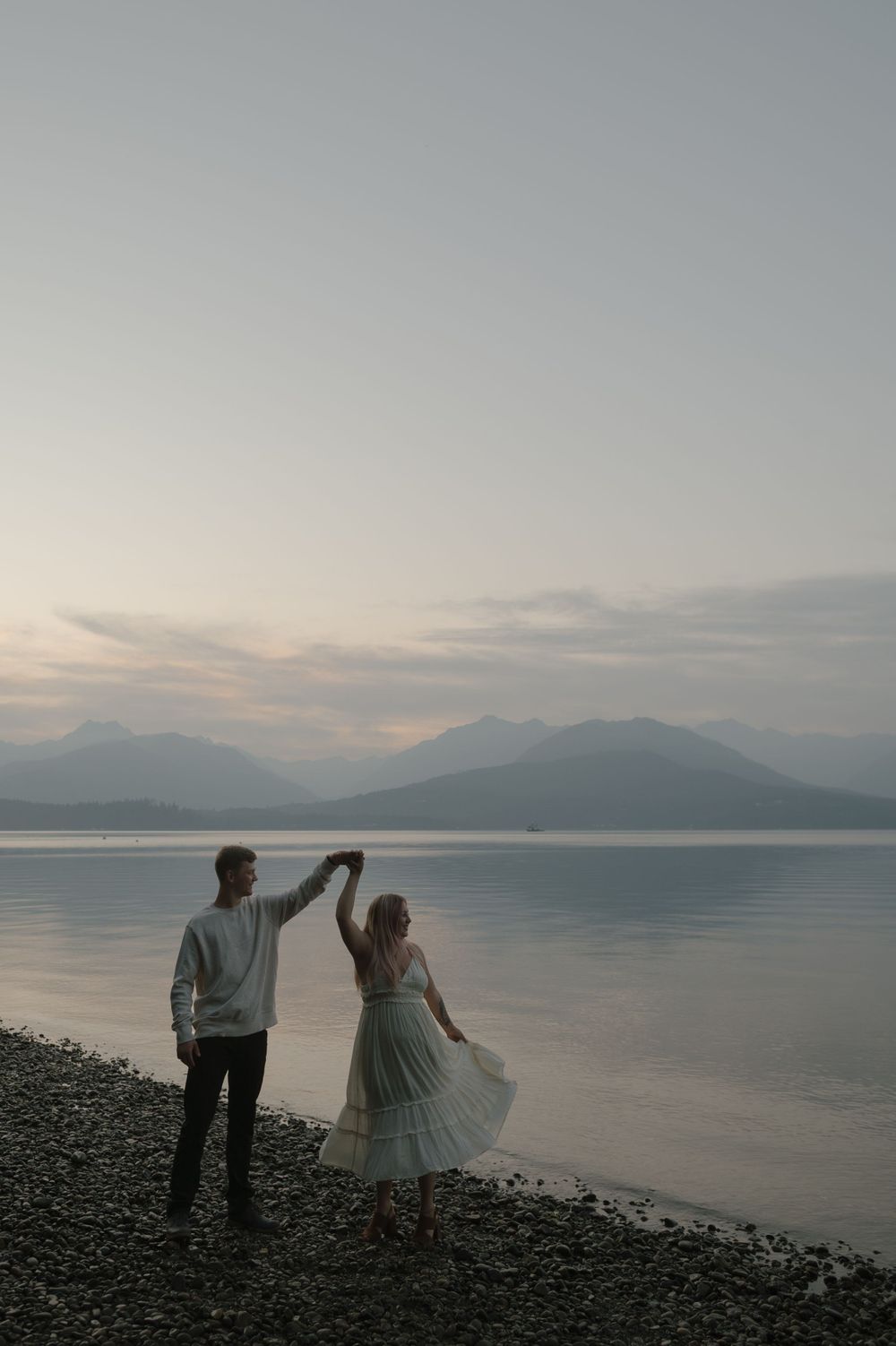 Couple dancing on a rocky beach at sunset with mountains and calm water in the background.