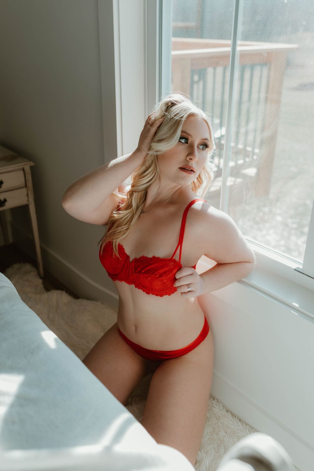 Woman in red lace lingerie by window with natural sunlight streaming in.