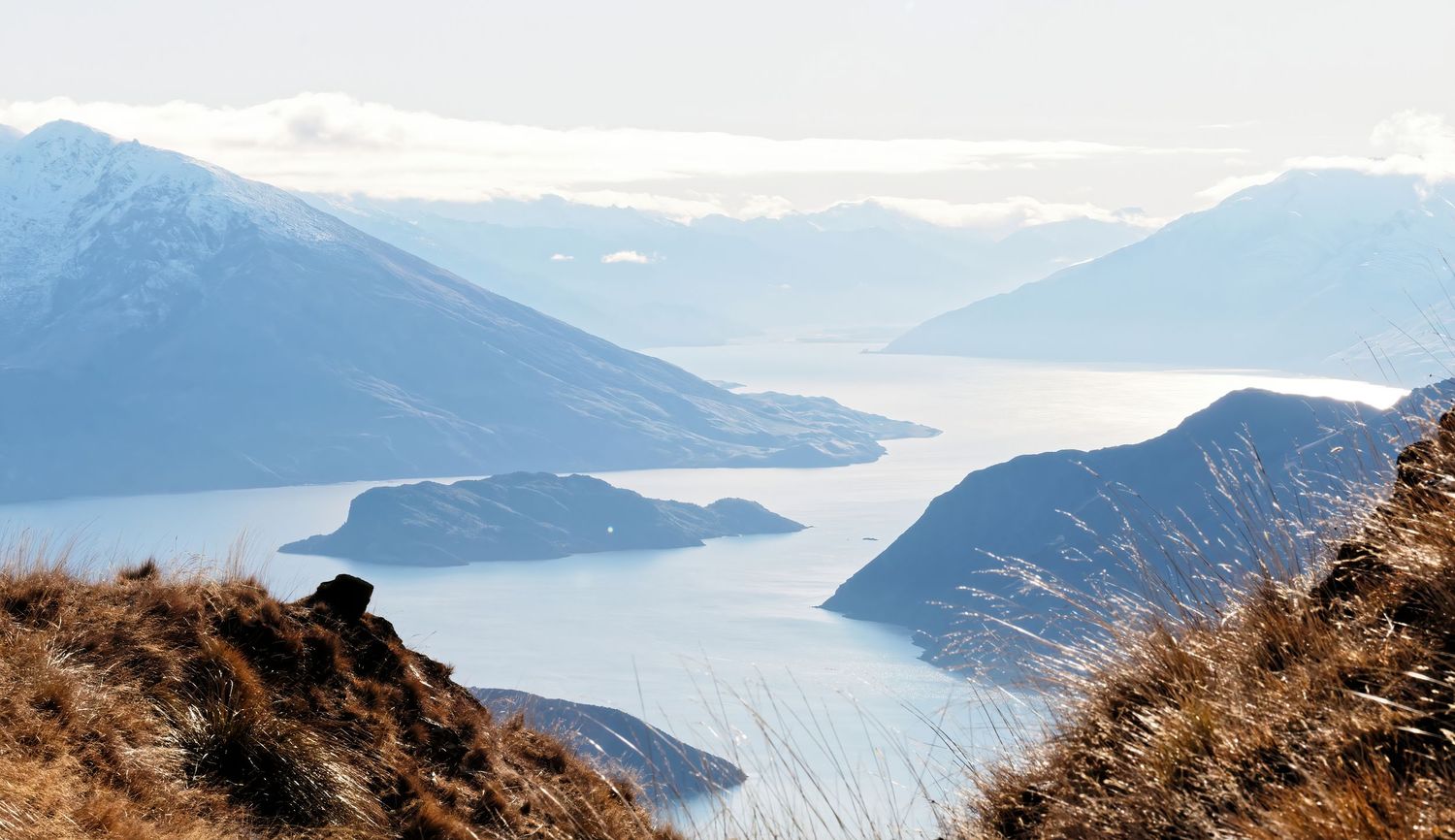 Scenic mountain landscape overlooking a winding lake or fjord with snow-capped peaks in the distance.