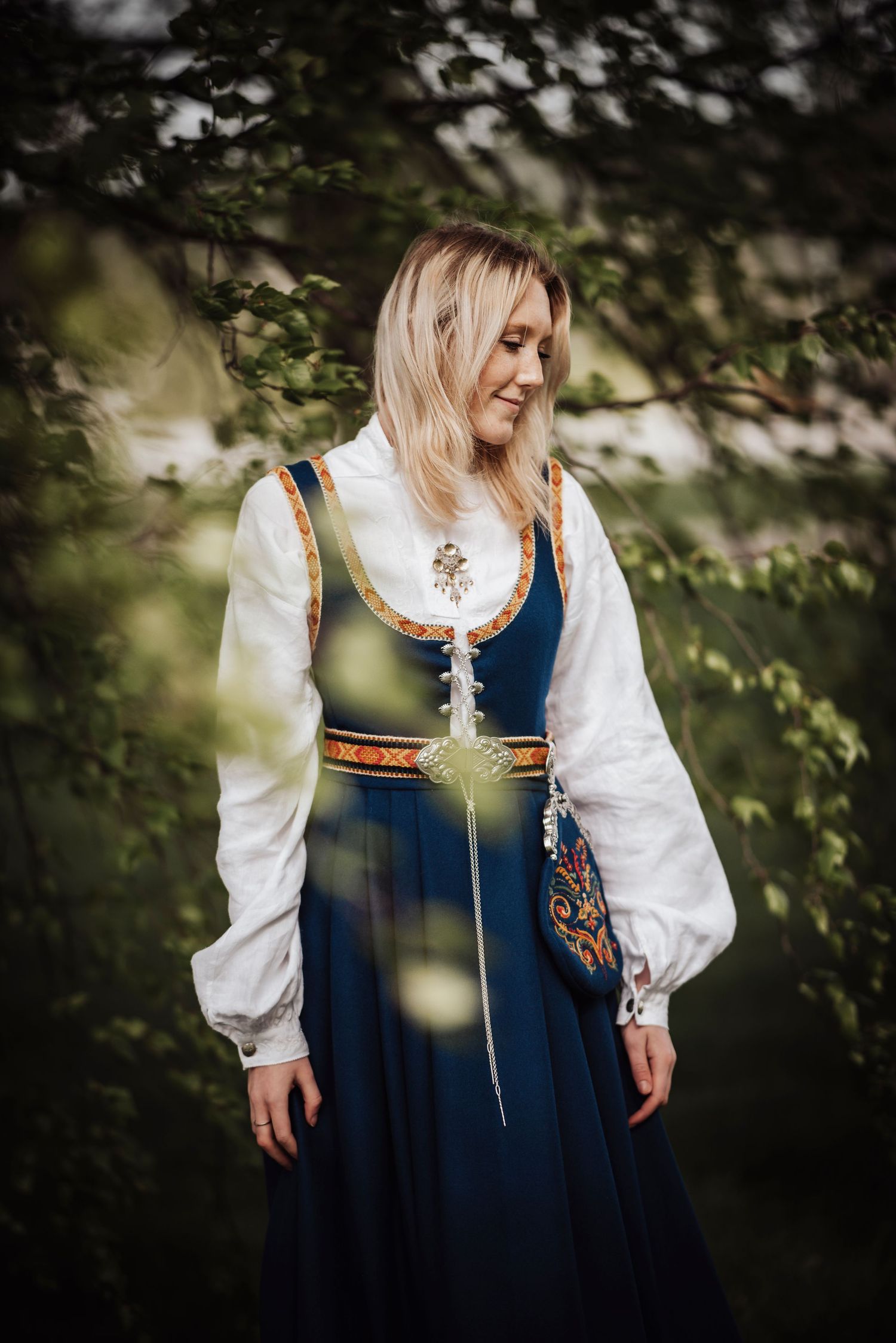 Norwegian traditional costume with navy blue dress and white puffy sleeves shown against leafy natural backdrop.