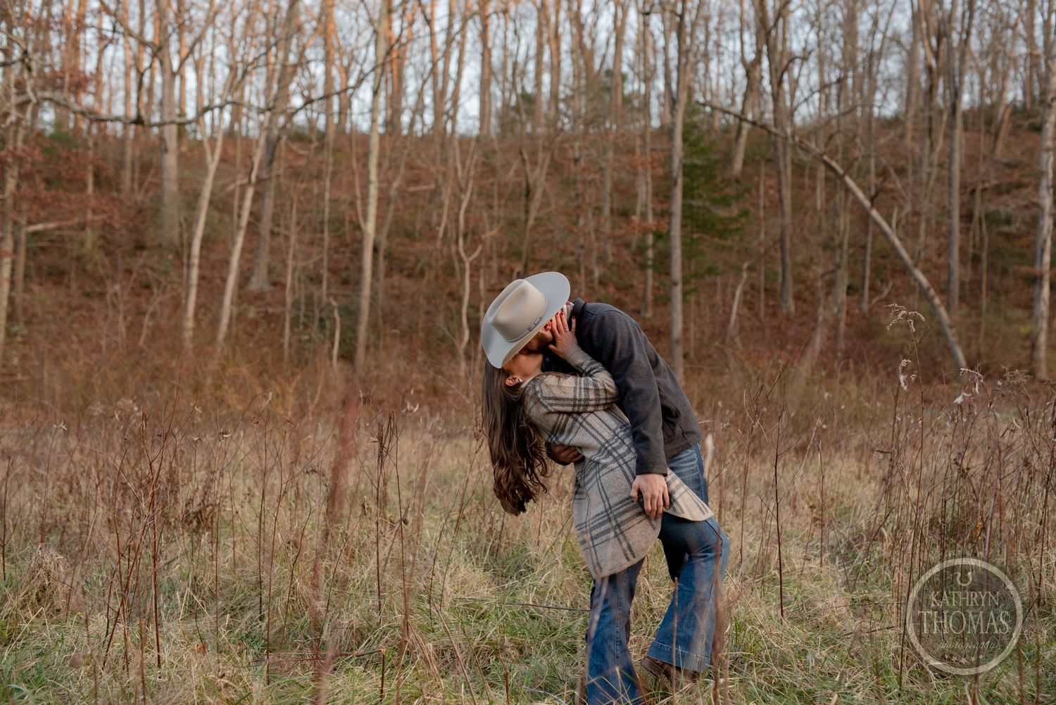 A romantic couple embraces in a fall field surrounded by bare trees and brown grass.