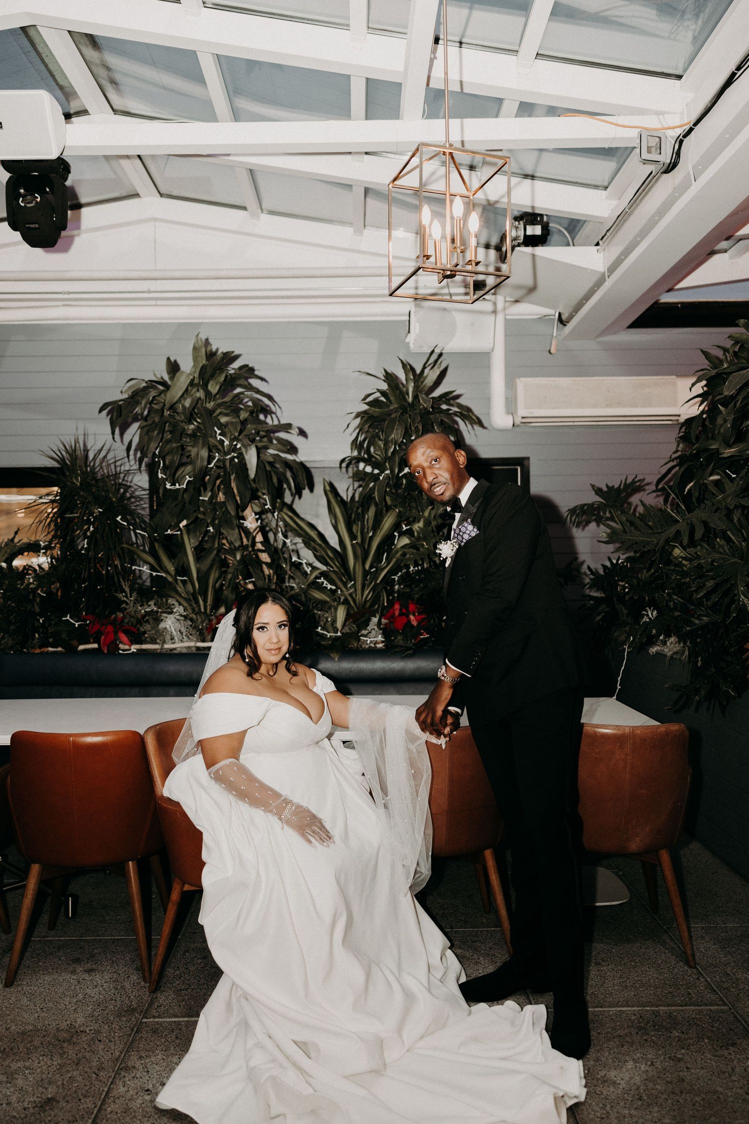 Bride sits beside groom in black tuxedo at elegant indoor wedding venue with lush greenery. Providence RI Miranda Photo & Co