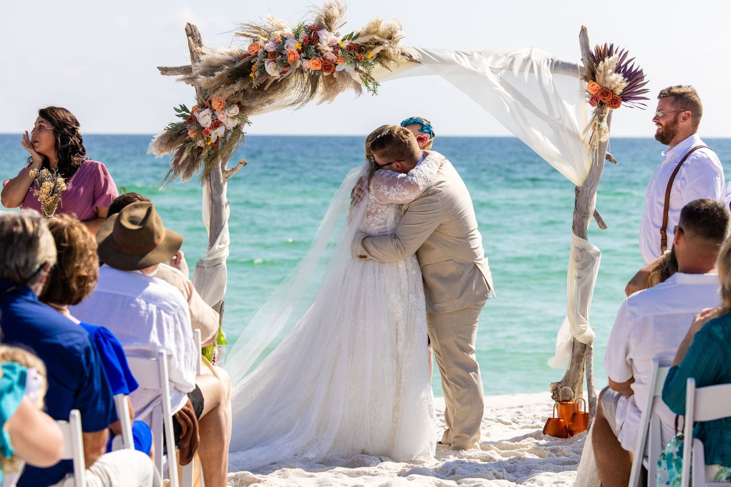 Beach wedding ceremony under wooden arch decorated with flowers and white fabric.