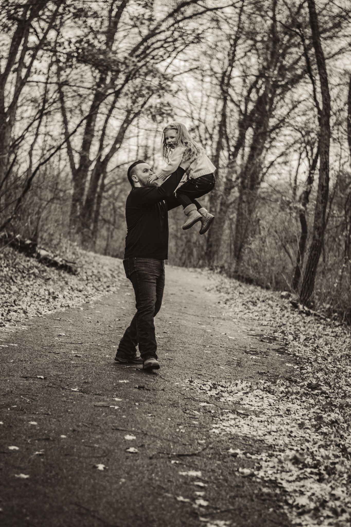 Playful scene of lifting and twirling on an autumn forest path lined with bare trees and fallen leaves.