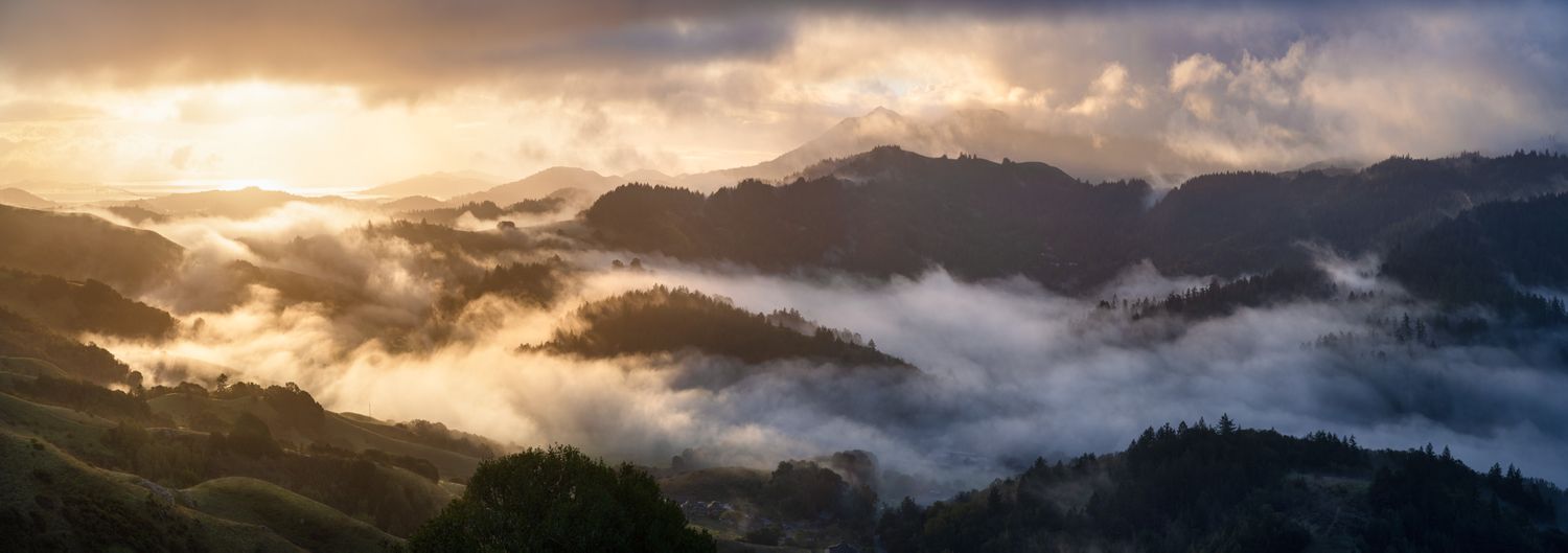 A stunning panoramic view of misty mountain peaks at sunrise with dramatic clouds and golden light filtering through.