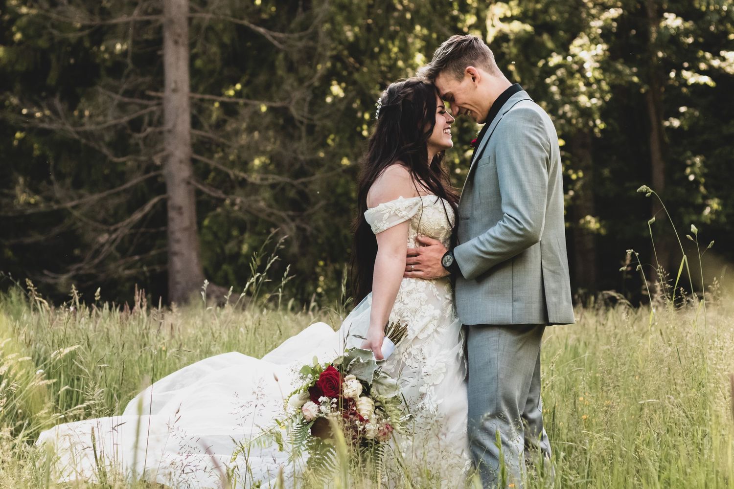 Intimate moment between newlyweds standing in sunny meadow with forest background.