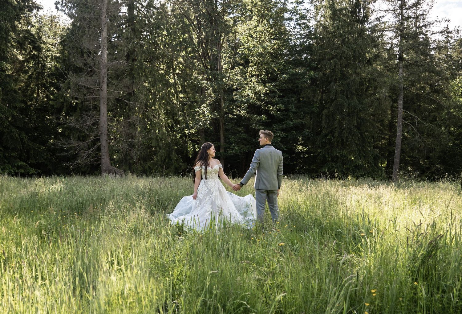 Couple walks through tall grass meadow in wedding attire with evergreen forest backdrop.