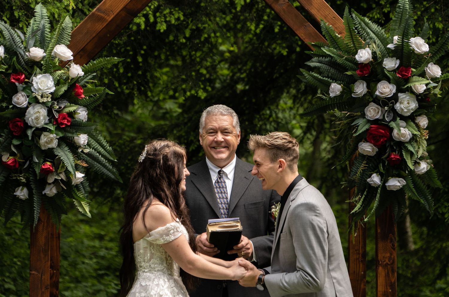 Outdoor wedding ceremony under wooden arch decorated with red and white floral arrangements.