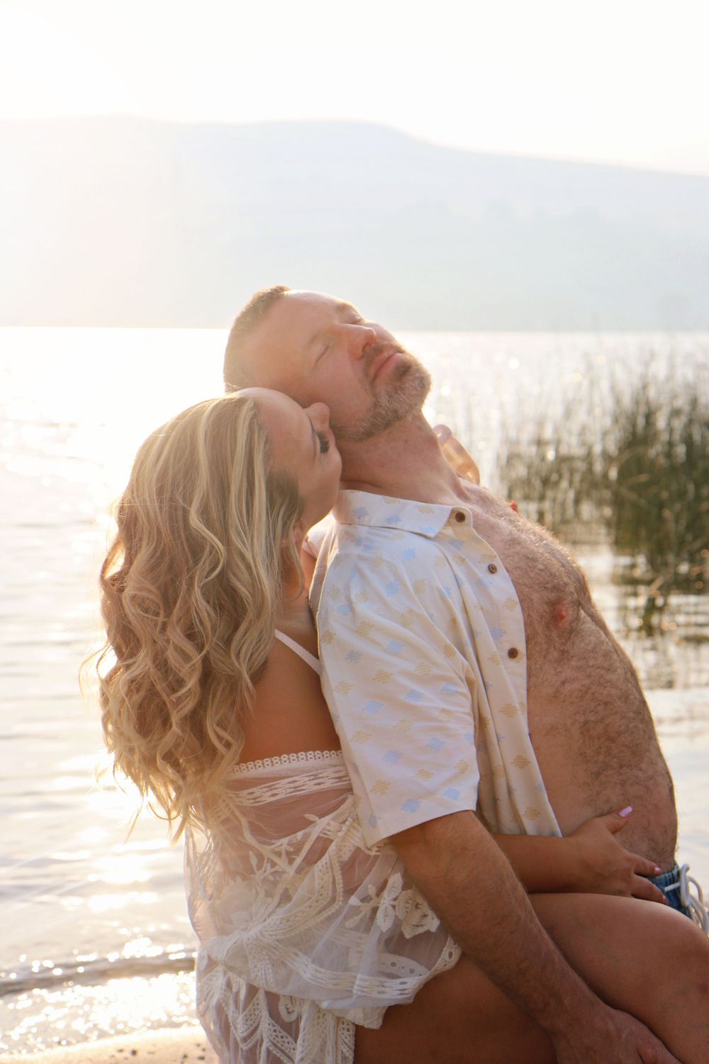 Tender waterfront embrace between couple in white clothing during sunset.