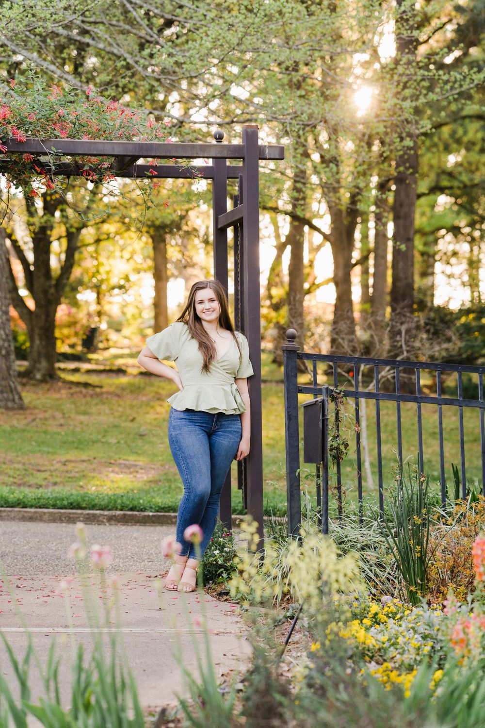 Woman in wrap blouse leans against garden archway during golden hour with flowers blooming.