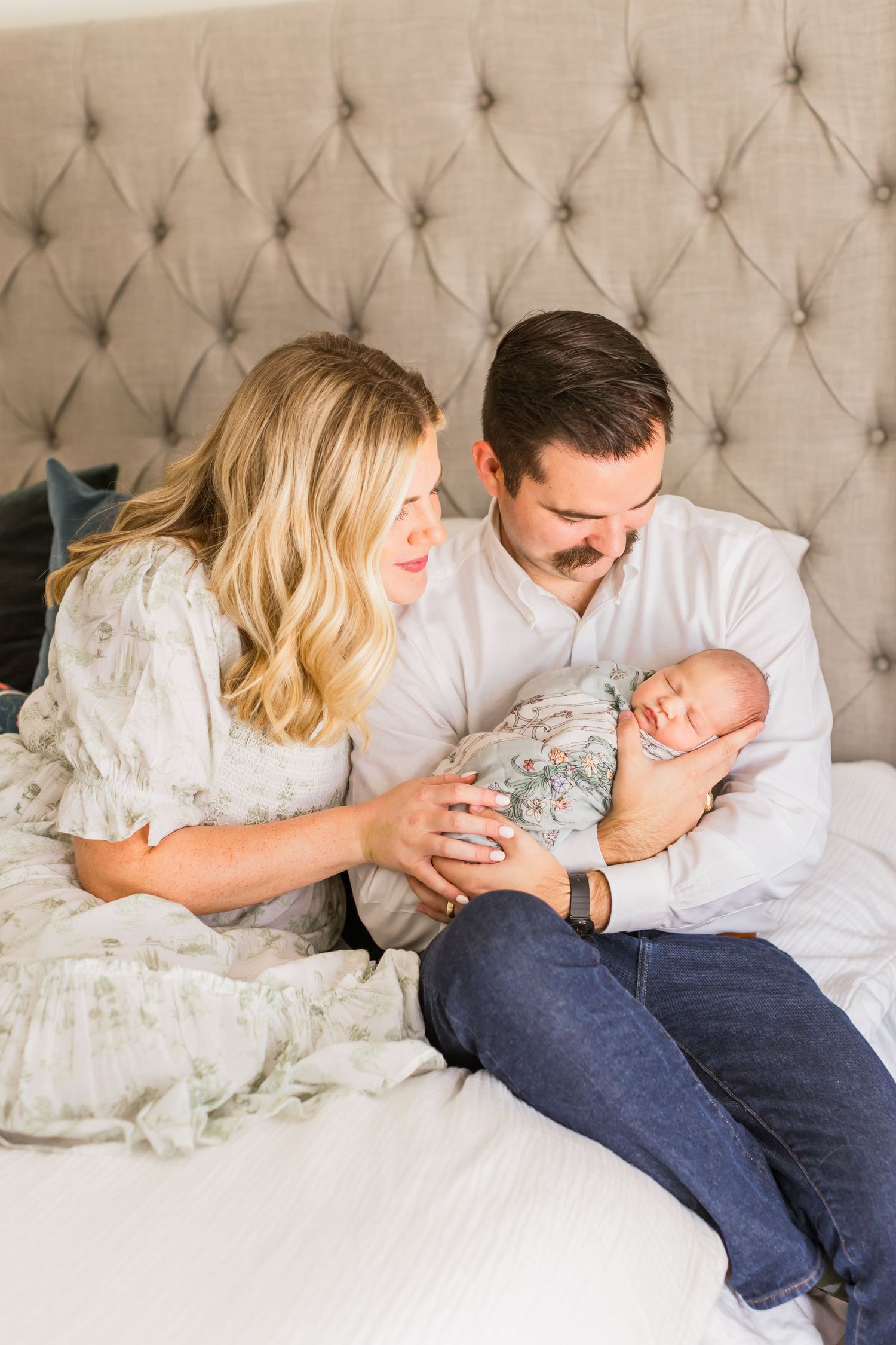 Family of three sharing intimate moment on bed with sleeping newborn in floral wrap.