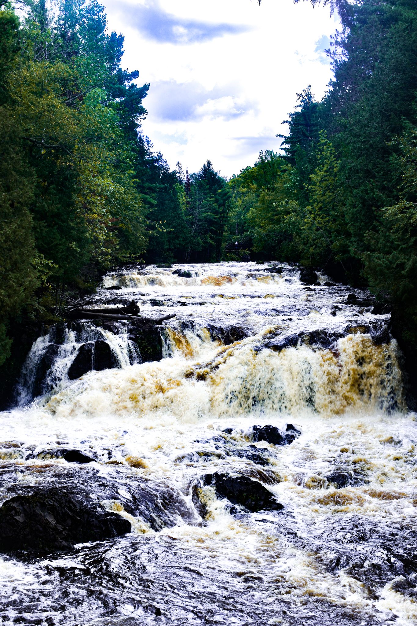 Powerful rushing waterfall cascades over rocky terrain through lush forest under cloudy skies.