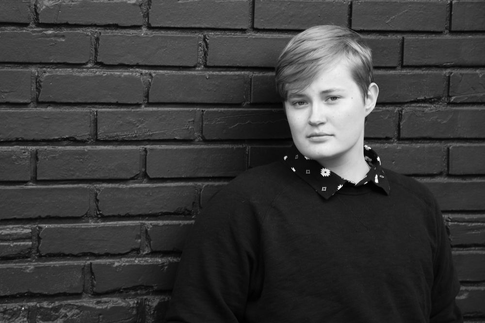 Black and white portrait of a graduate leaning against a black brick wall. face is looking away but eyes on camera.