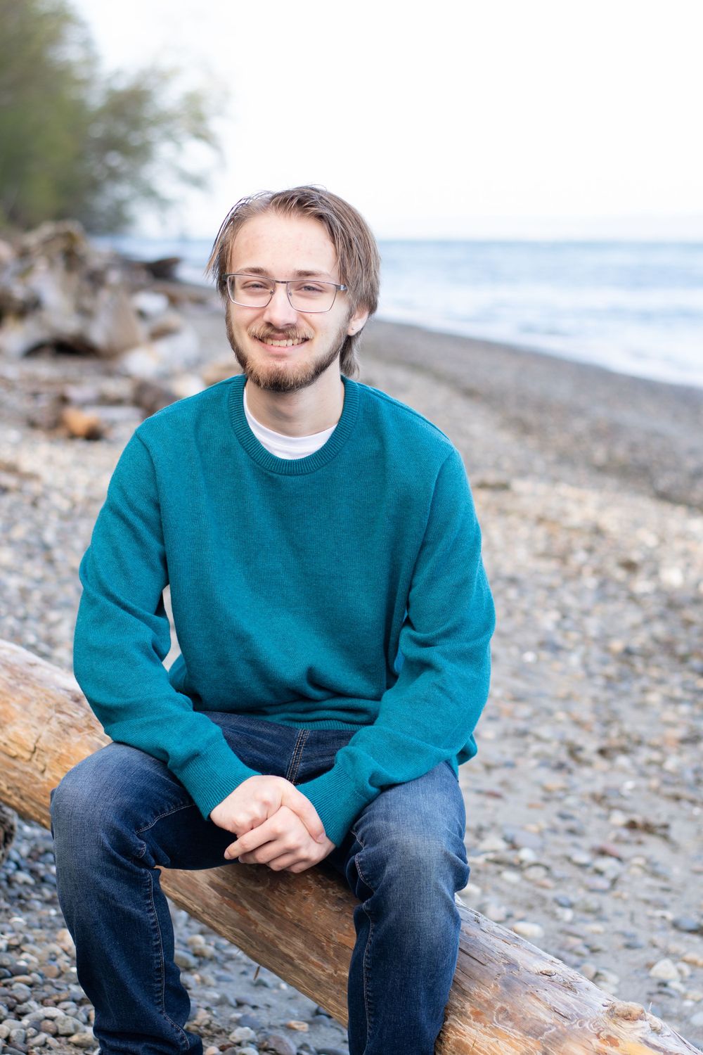 graduate faces the camera as he sits at the beach on a log framed by trees and the Puget Sound.