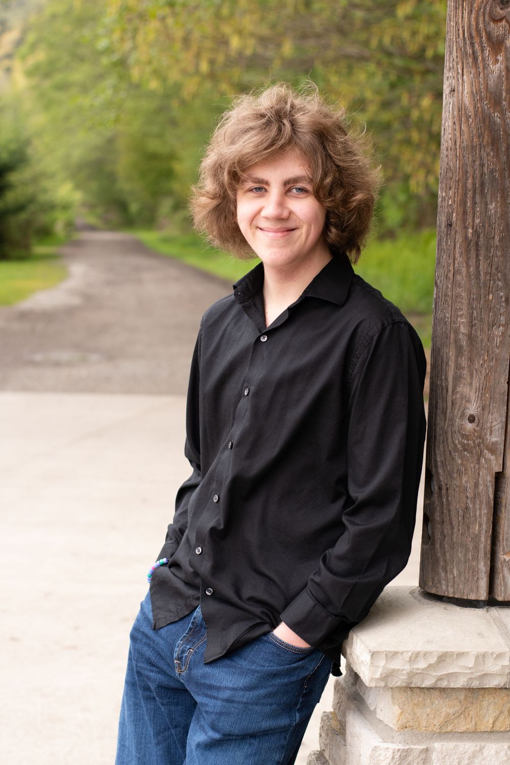 male graduate leaning against a pole with a tree lined trail stretches out behind him to his right.