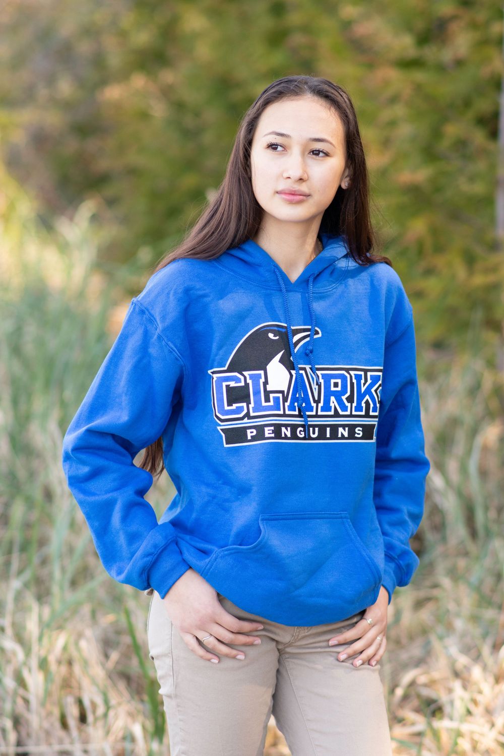 female graduate wearing a sweatshirt from her declared college. she is looking away and framed by beach grass and trees.
