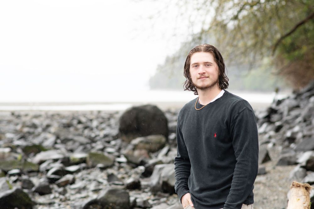male graduate stands on rocky PNW shore framed by foggy trees and water.