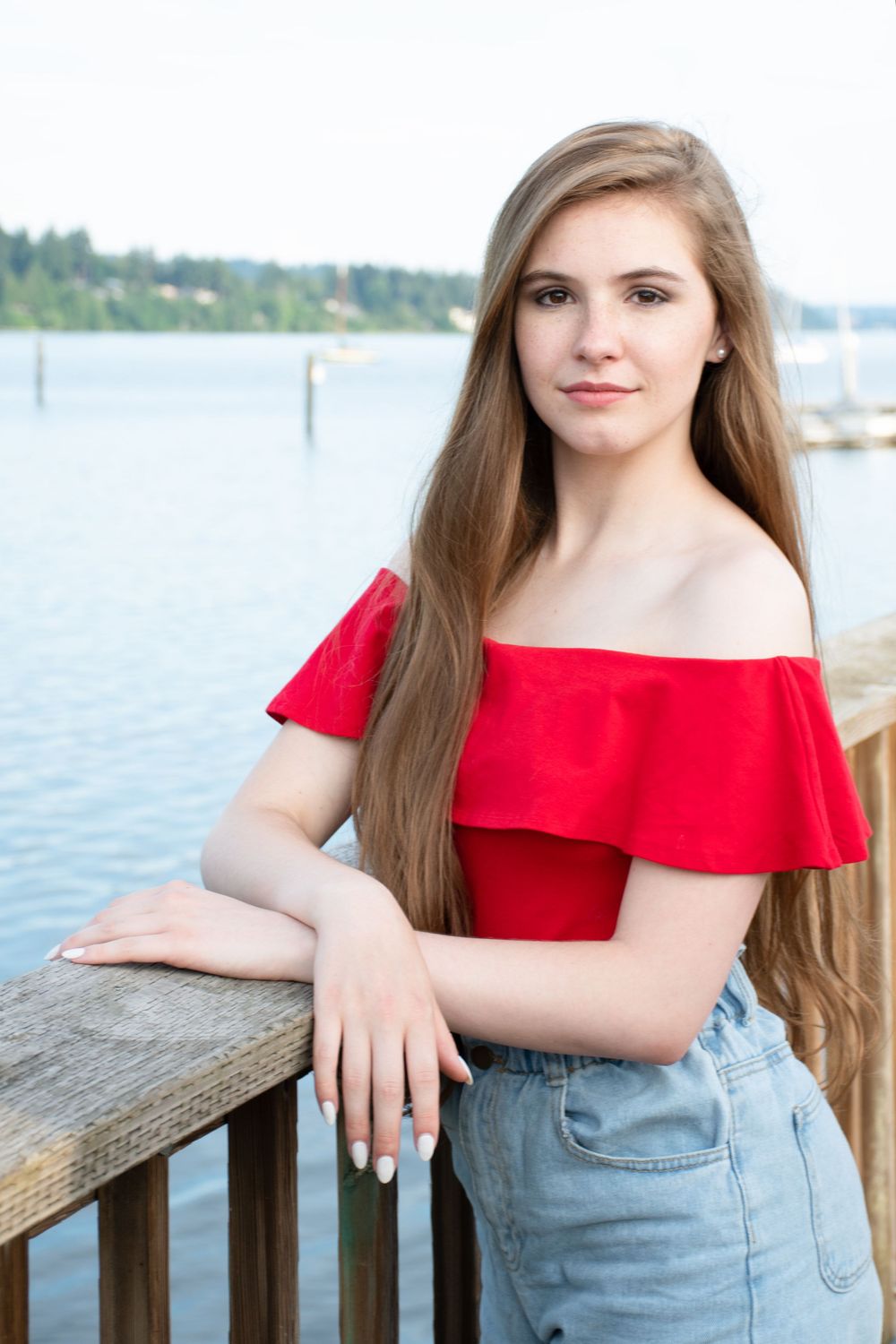 female graduate stand on the pier with glassy blue water and opposing shore framing her as she smiles slightly.