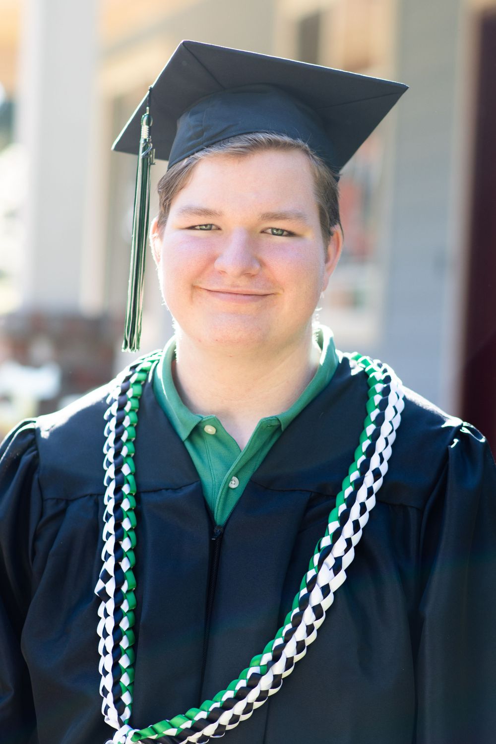 male graduate stands in cap and gown with celebratory ties. He has a slight smile while standing in front of a porch.