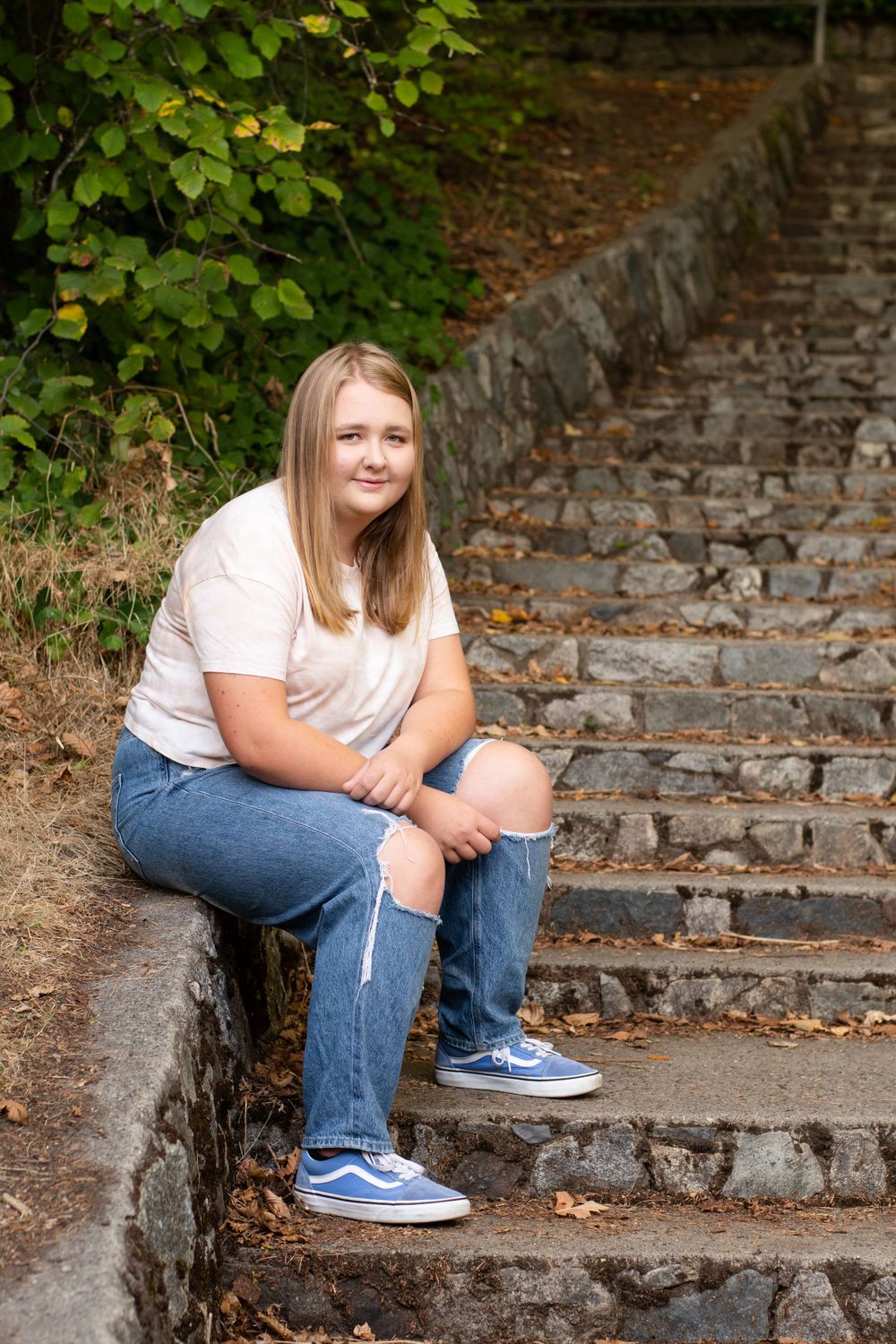 graduate sits on the edge of mossy stairs framed by deep green tress and bushes. long blond hair and a slight smile.