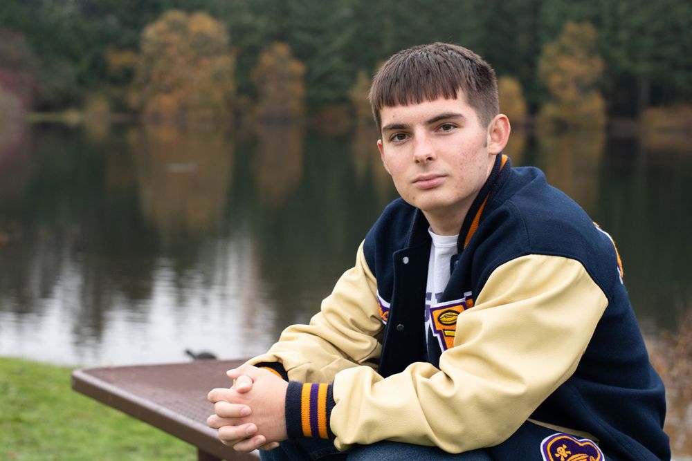 Male graduate sits on picnic table wearing his letterman jacket framed by the glassy lake and fall leaves.