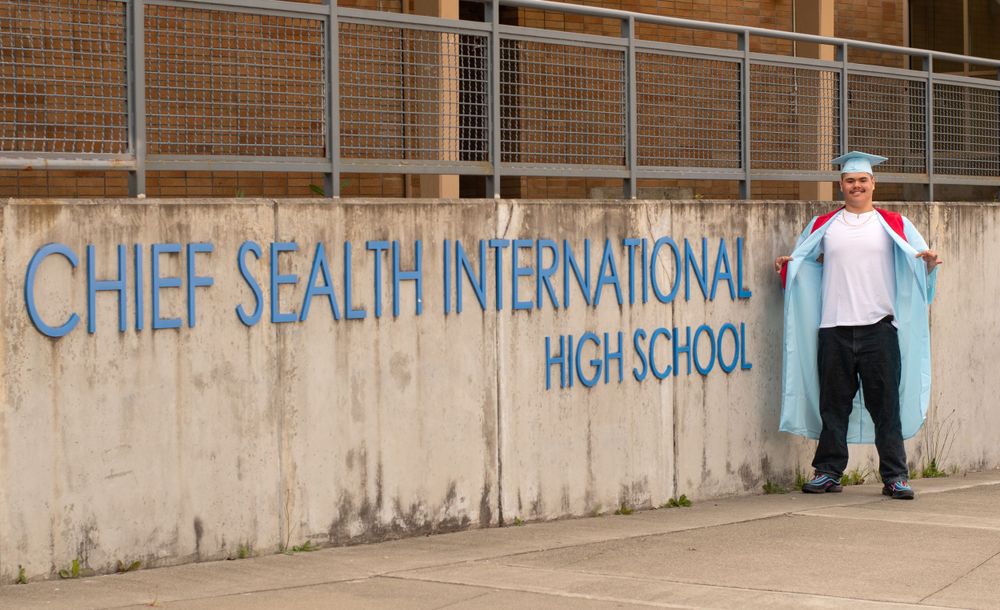 grad puffs his chest in cap and gown while standing next to the name of his school.