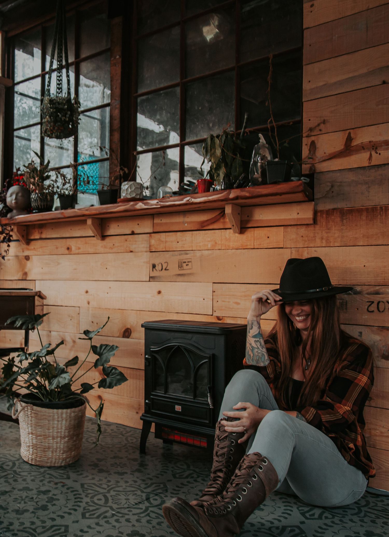 Smiling woman in a black hat and plaid flannel sits on a rustic wooden porch beside a small black stove and potted plants.