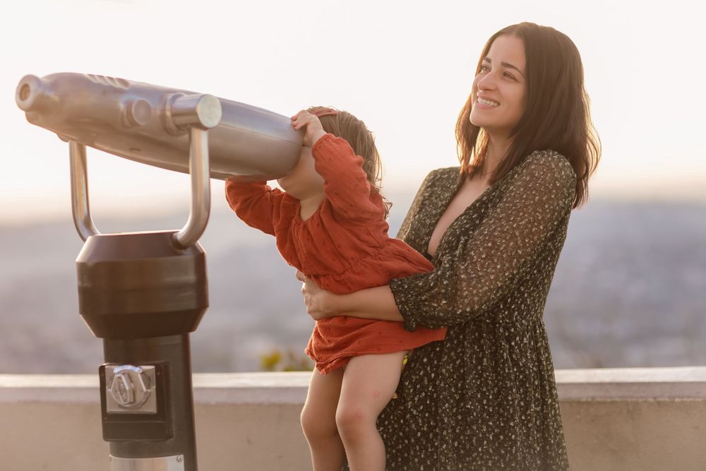 A child explores mounted binoculars at an observation deck while being held up by an adult in a patterned dress.