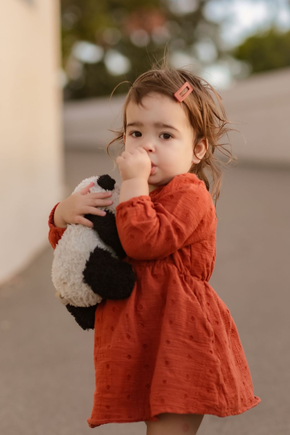 A small child in a red dress hugs a stuffed panda toy while wearing hair clips.