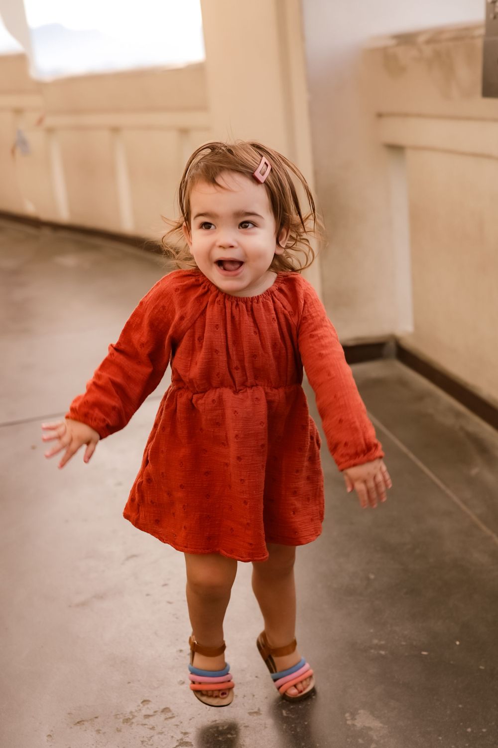 A joyful toddler in a red dress and colorful socks laughs while walking indoors.