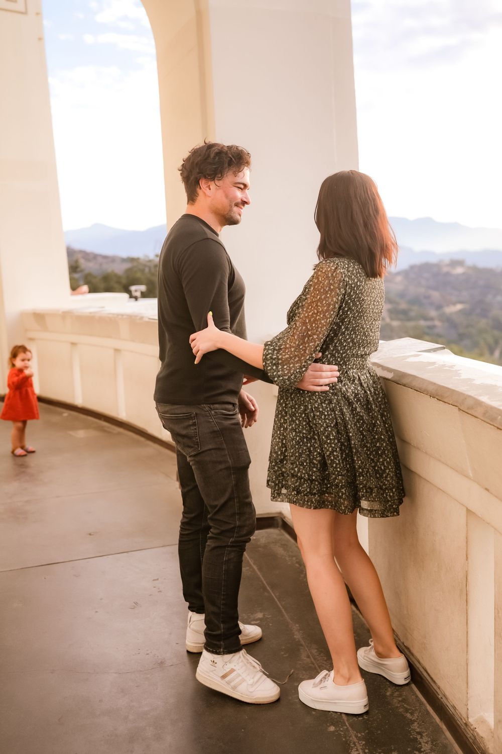 A couple shares a loving moment on a balcony while a child in red plays in the background.