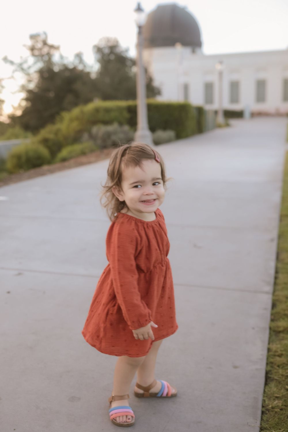 A toddler in an orange dress smiles while walking on a path near a domed building.