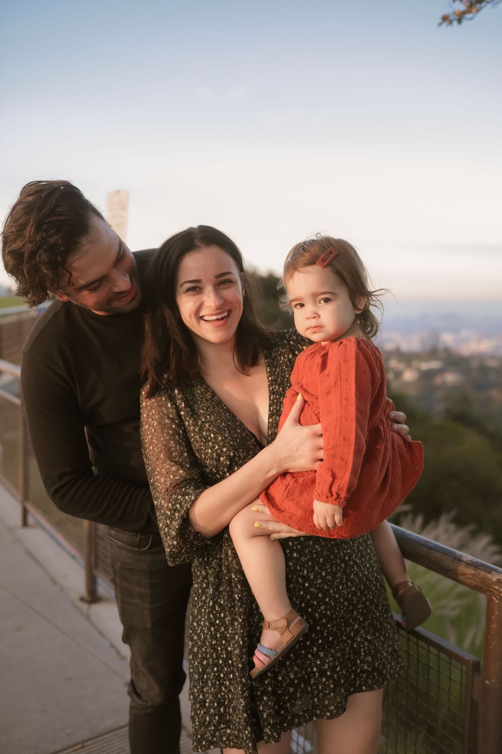 A family poses together on an outdoor terrace at sunset, with the parent in black holding a toddler in orange.