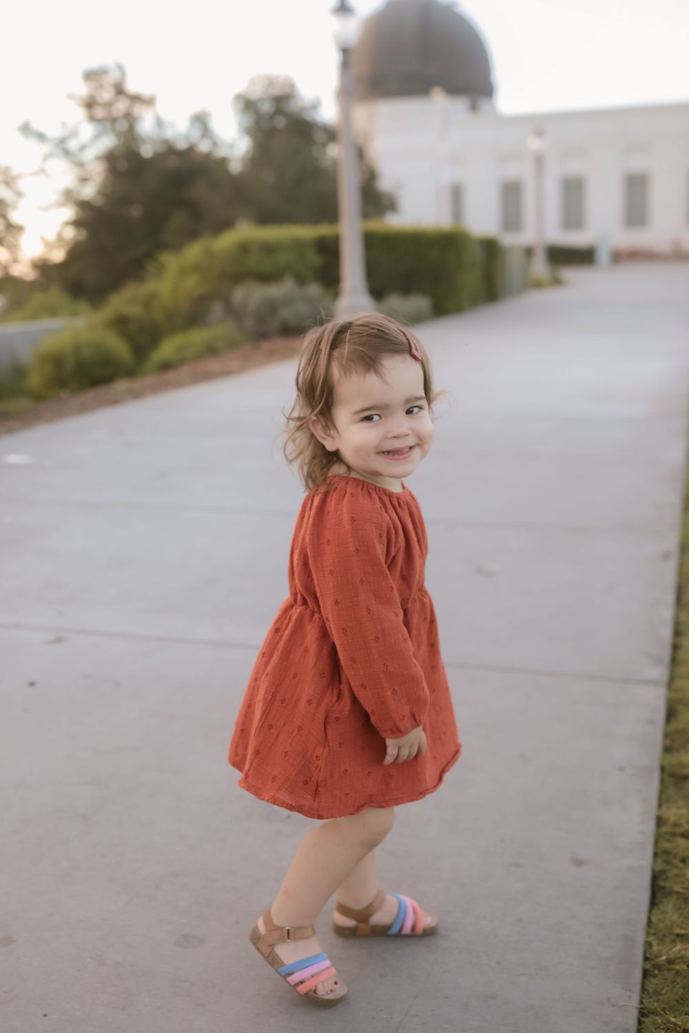 A small child in a coral dress poses playfully on a walkway near a domed structure.