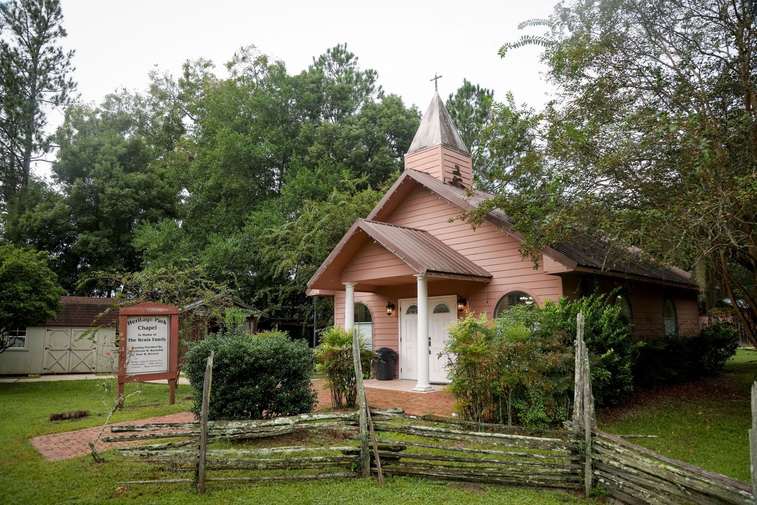 A charming rustic wooden chapel with steeple nestled among green trees and split-rail fence.
