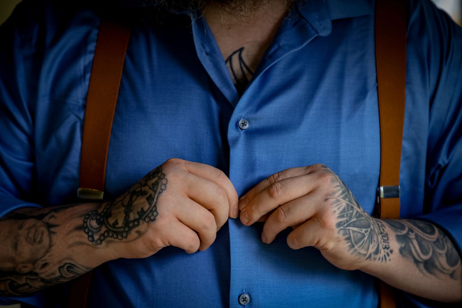Close-up of tattooed hands buttoning a blue dress shirt with brown leather suspenders.