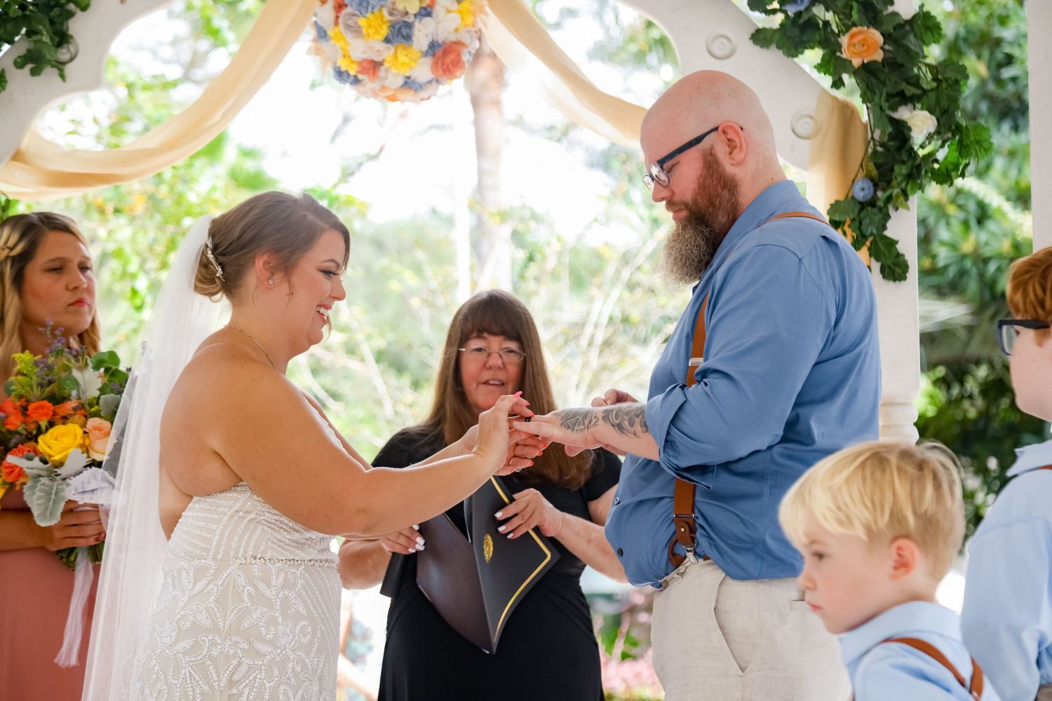 An outdoor wedding ceremony under a floral arch as rings are exchanged.