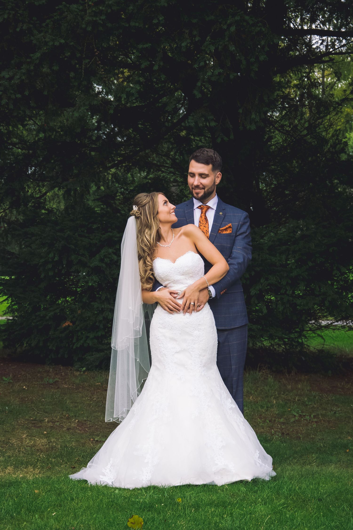 A wedding couple poses together on grass with lush greenery in the background.