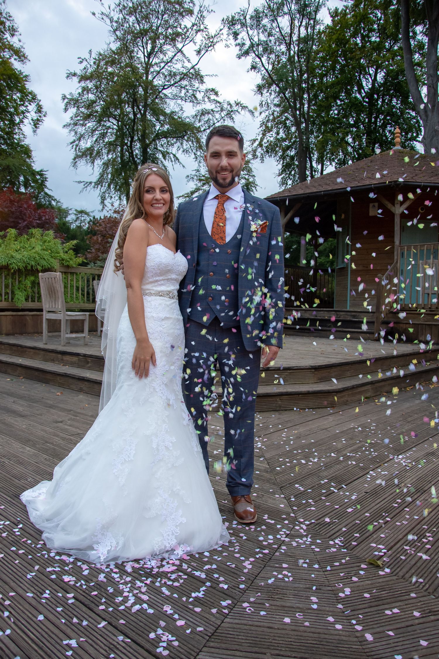 Newlyweds pose on wooden deck while confetti falls around them.