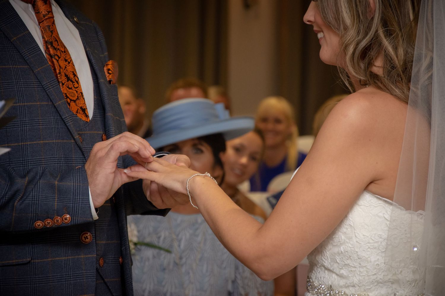 Close-up of a ring exchange during a wedding ceremony with guests visible in the background.