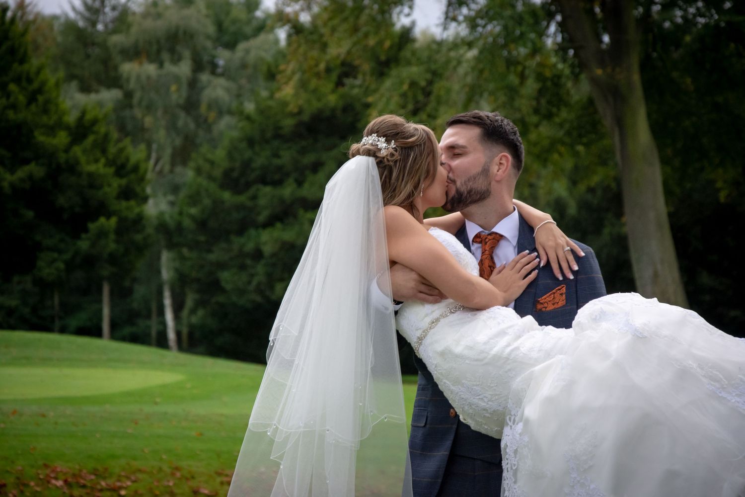 Groom carries bride in white dress during outdoor wedding photoshoot.