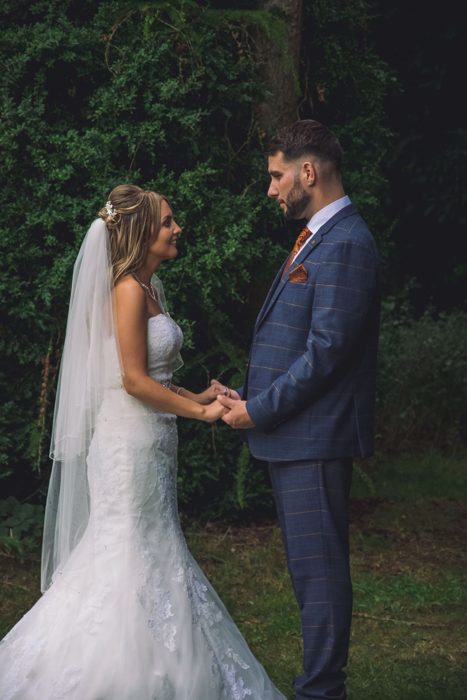 Bride and groom hold hands while standing on grass with trees in the background.