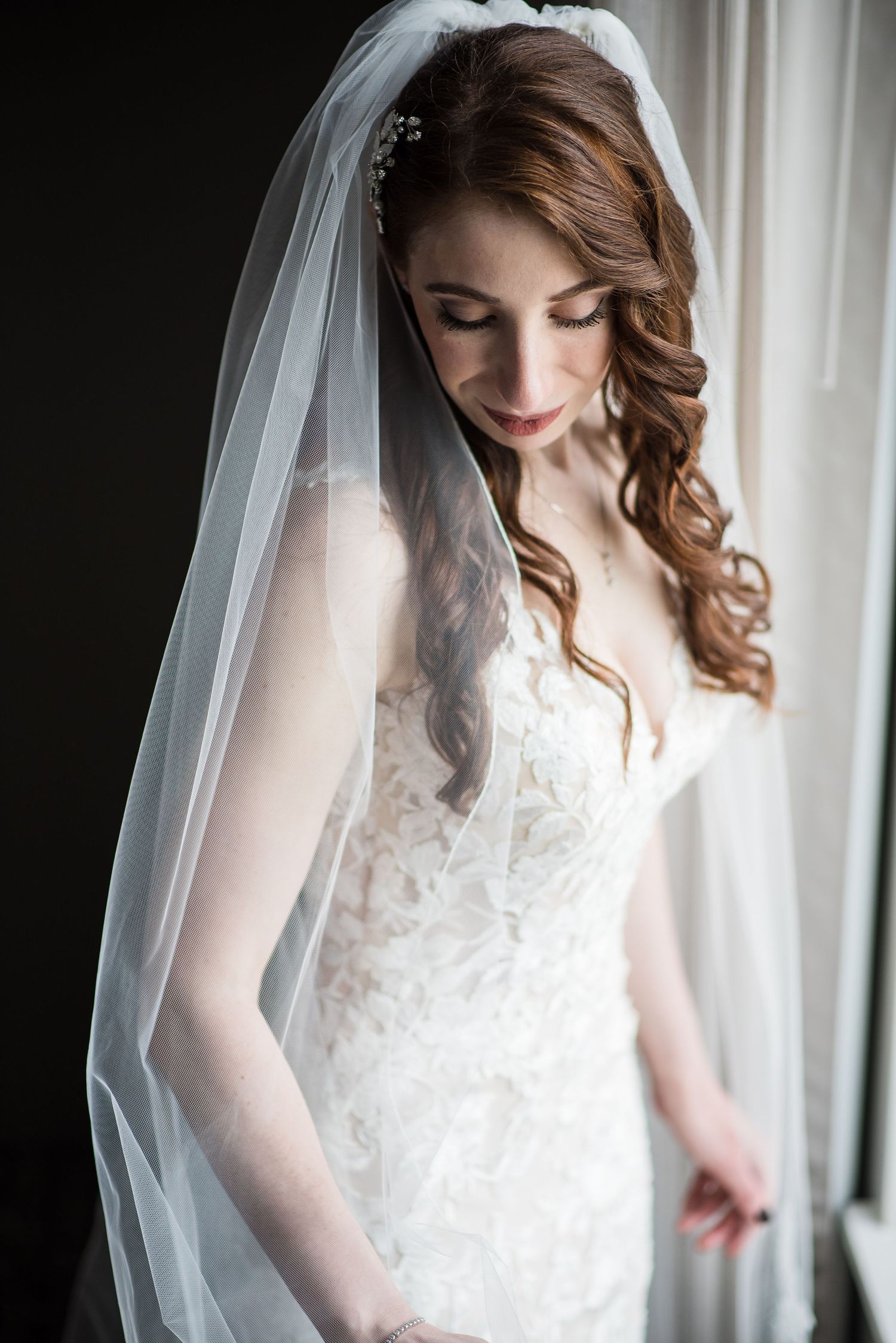 Elegant bride in lace wedding gown and veil gazing down by a window on her wedding day.