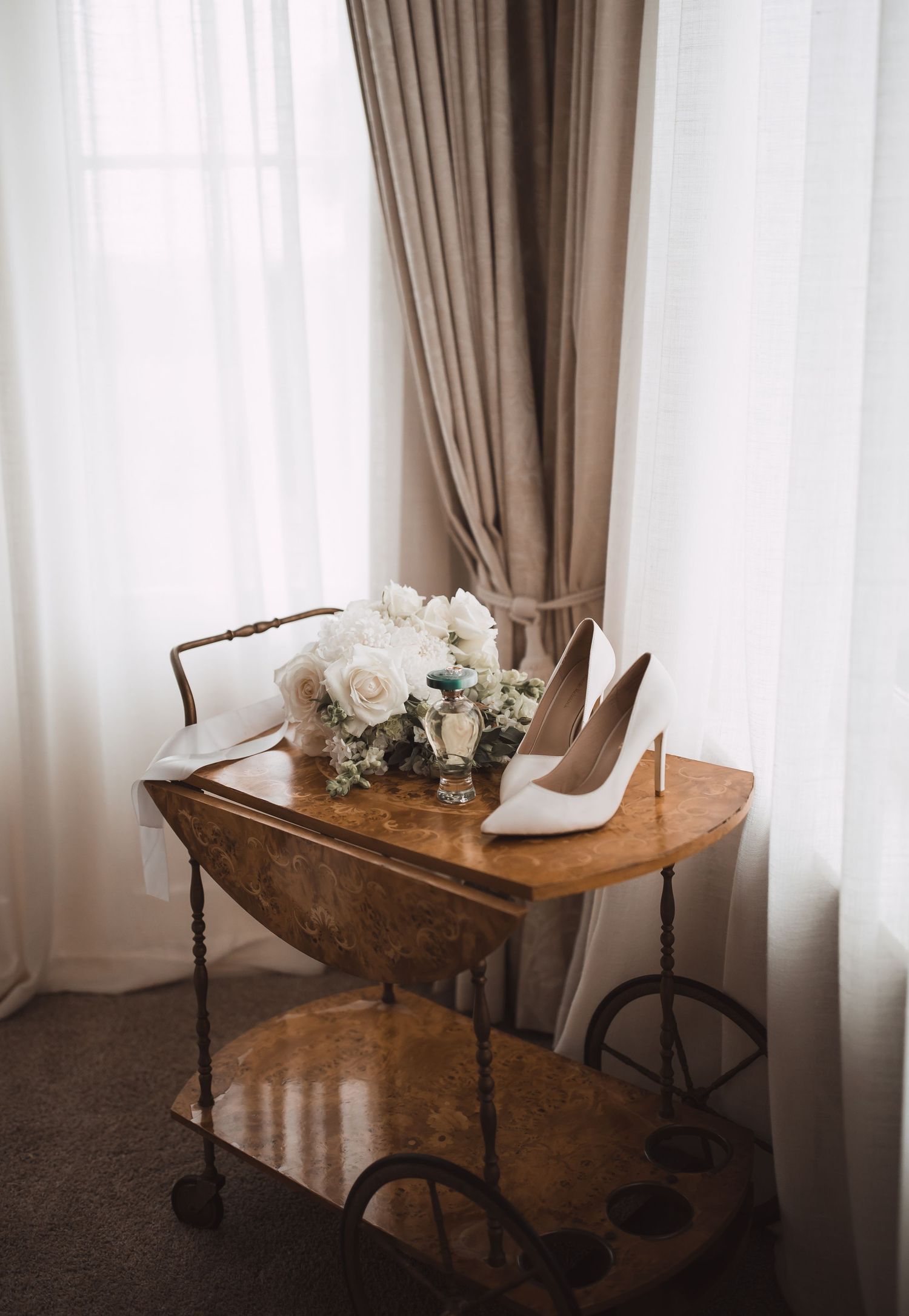 Minimalist wedding setup with wooden table, white chairs and floral arrangements by window.